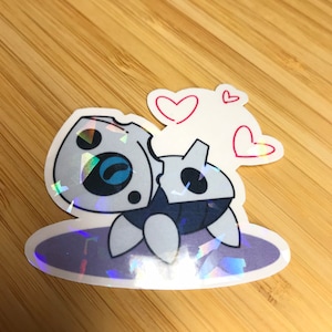May include: A holographic sticker of a cartoon robot with a cracked shell, surrounded by three red hearts. The robot is on a holographic background.