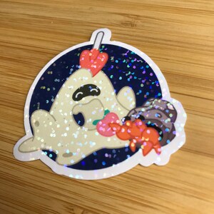 May include: A glittery sticker featuring a cartoon ghost character with a black mask, holding a strawberry and a crab. The ghost has a strawberry on its head. The sticker has a dark blue background with glitter.