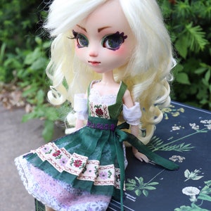 May include: A doll with blonde hair wearing a green and white dirndl dress with a pink and white petticoat. The doll is sitting on a black box with a floral pattern.