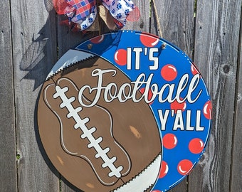 May include: A round wooden door hanger with a painted football design and the words "It's Football Y'all". The football is brown with white laces and a white outline. The background is blue with red polka dots. A red, white, and blue bow is attached to the top of the door hanger.