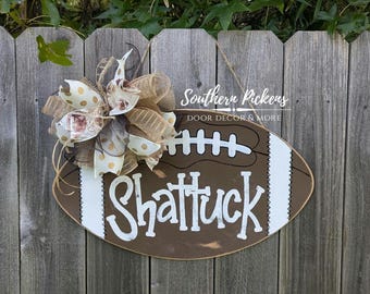 May include: A brown and white football-shaped door hanger with the name "Shattuck" written in white paint. A brown, white, and gold bow is attached to the top of the hanger. The hanger is hanging on a wooden fence. The text "Southern Pickers Door Decor & More" is visible in the top right corner of the image.
