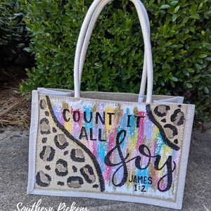 May include: A rectangular tote bag with white handles and a beige burlap body. The bag features a colorful painted design with the words "COUNT IT ALL JOY" and "JAMES 1:2" in black script. Leopard print accents the design.