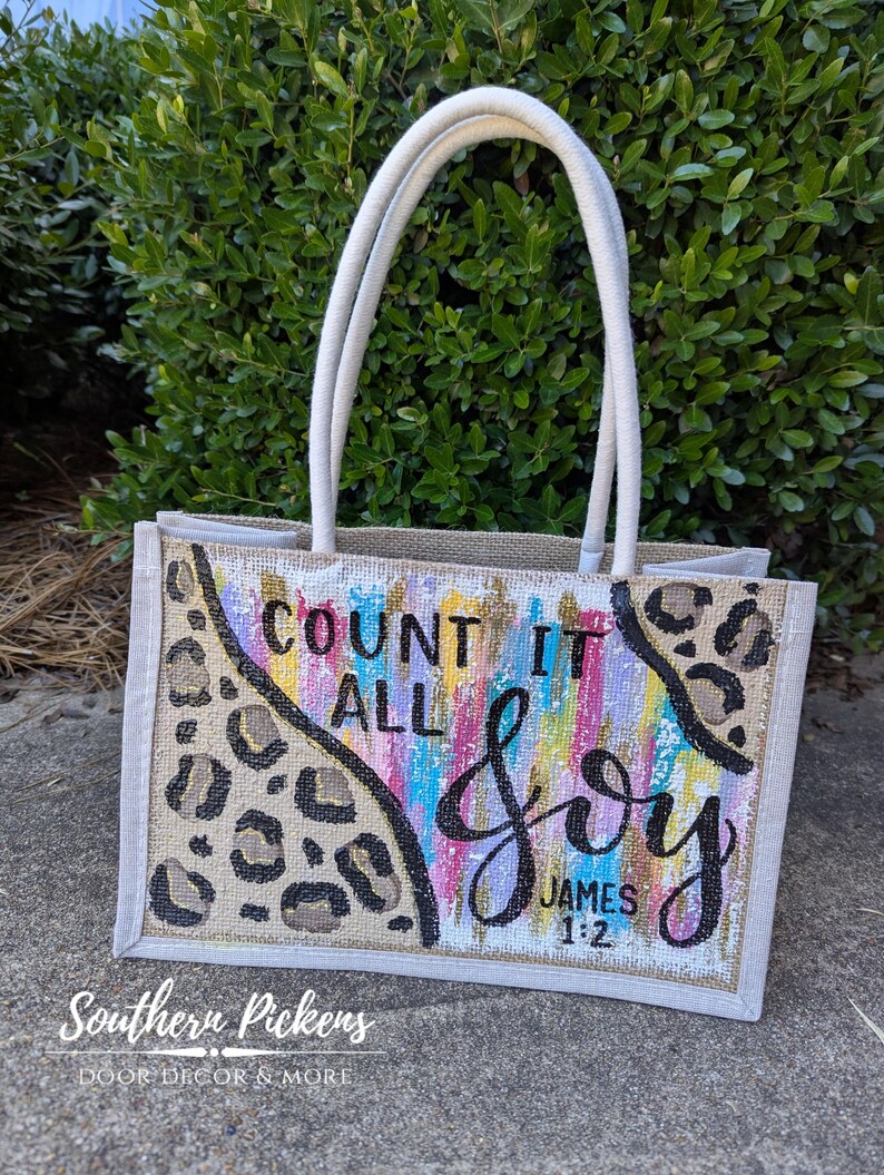 May include: A rectangular tote bag with white handles and a beige burlap body. The bag features a colorful painted design with the words "COUNT IT ALL JOY" and "JAMES 1:2" in black script. Leopard print accents the design.