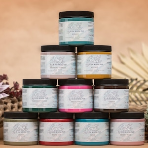 May include: A stack of ten 8 oz. jars of Silk All-in-One Mineral Paint in various colors, including Midnight Green, Prickly Pear, Lavender, and Desert Rose. The jars are arranged in a pyramid shape on a brown surface.