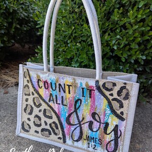 May include: A beige burlap tote bag with cream-colored handles. The front features the words "Count It All Joy" in black script, with colorful vertical brushstrokes and leopard print accents. The text "James 1:2" is also visible.