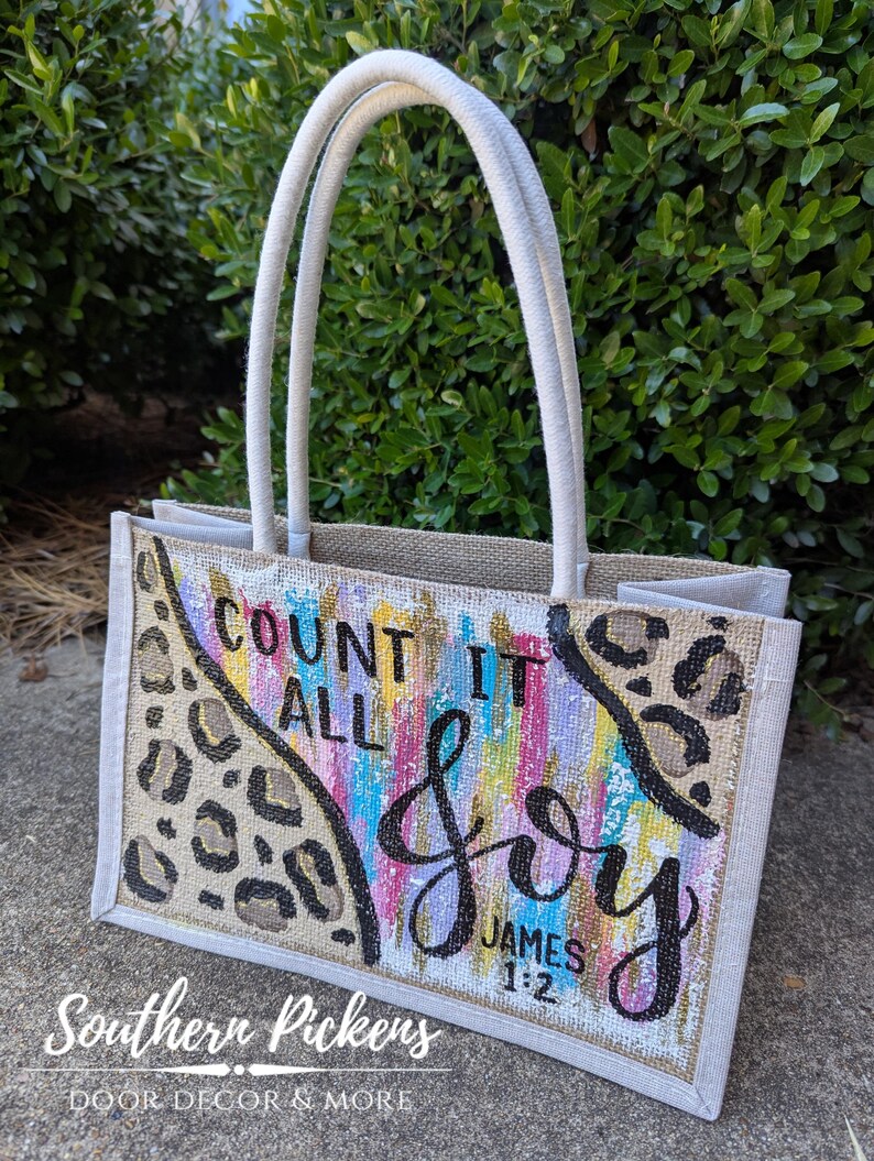 May include: A beige burlap tote bag with cream-colored handles. The front features the words "Count It All Joy" in black script, with colorful vertical brushstrokes and leopard print accents. The text "James 1:2" is also visible.