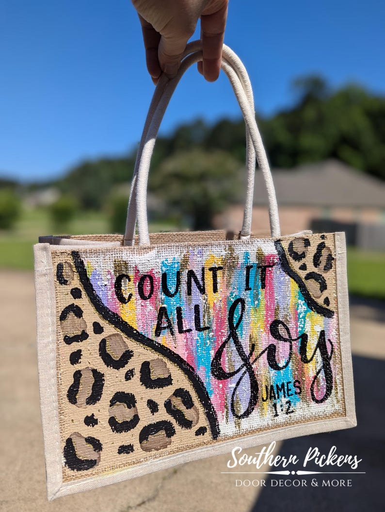 May include: A rectangular burlap tote bag with a cream-colored handle. The bag features a leopard print design and the words "COUNT IT ALL JOY" in black script with colorful brushstrokes. The text "JAMES 1:2" is also visible.