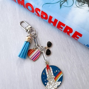 Op de afbeelding: Een zilveren sleutelhanger met een blauwe kwast, een hartvormige bedel met regenboogstrepen en een bedel met een zwarte zonnebril. De hoofdbedel is een blauwe cirkel met een raket, regenboog en sterren.