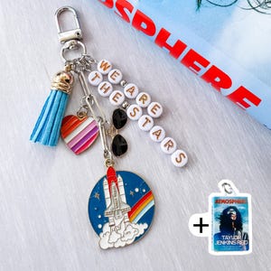 Op de afbeelding: Een zilveren sleutelhanger met een blauwe kwast, een hart met regenboogstrepen en een bedel met een raket die over een regenboog lanceert. De sleutelhanger heeft ook de tekst "WE ARE THE STARS" en een klein label met de tekst "ATMOSPHERE".