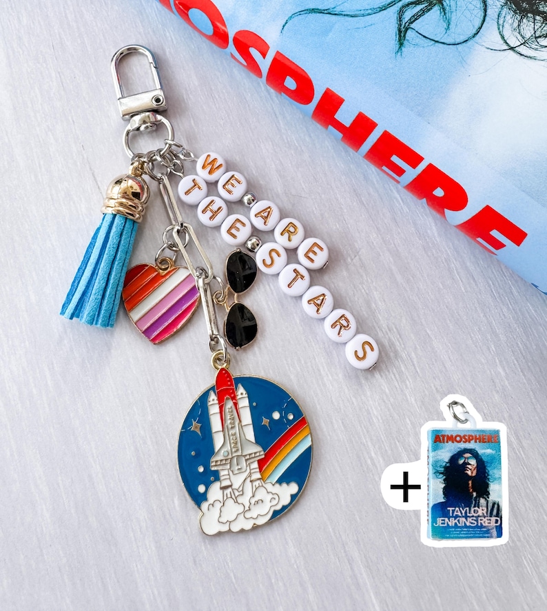 Op de afbeelding: Een zilveren sleutelhanger met een blauwe kwast, een hart met regenboogstrepen en een bedel met een raket die over een regenboog lanceert. De sleutelhanger heeft ook de tekst "WE ARE THE STARS" en een klein label met de tekst "ATMOSPHERE".