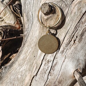 May include: A gold-coloured, round pet tag with a textured surface hangs from a gold-coloured ring. The tag is suspended from a small loop attached to the ring. The background is a weathered, light-coloured tree trunk.