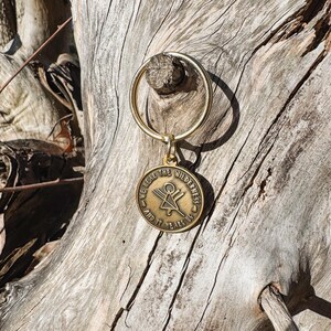 May include: A gold-coloured keyring with a circular pendant. The pendant features the text "WE HEAR THE WILDERNESS AND IT HEARS US" around a central design. The keyring is attached to a wooden surface.