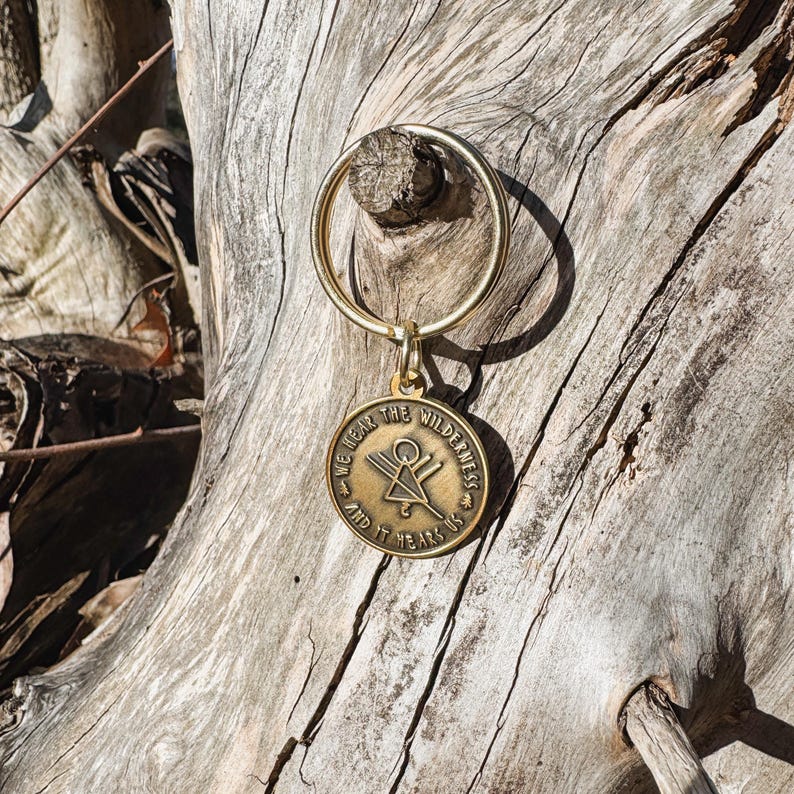 May include: A gold-coloured keyring with a circular pendant. The pendant features the text "WE HEAR THE WILDERNESS AND IT HEARS US" around a central design. The keyring is attached to a wooden surface.