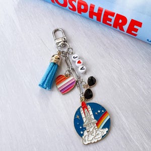 Op de afbeelding: Een zilveren sleutelhanger met een blauwe kwast, een hartvormige bedel met regenboogstrepen en een ronde bedel met een raket die opstijgt met een regenboog. De sleutelhanger heeft ook de letters "J♡V" op witte kralen.