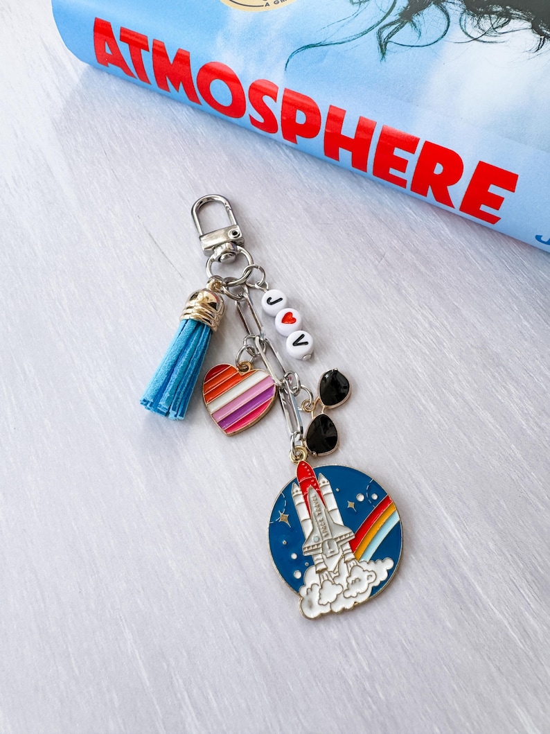 Op de afbeelding: Een zilveren sleutelhanger met een blauwe kwast, een hartvormige bedel met regenboogstrepen en een ronde bedel met een raket die opstijgt met een regenboog. De sleutelhanger heeft ook de letters "J♡V" op witte kralen.