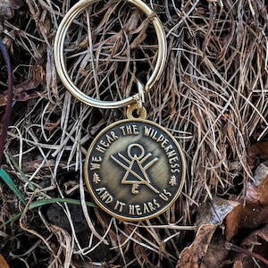 May include: A bronze-coloured keyring with a circular pendant. The pendant features the text "WE HEAR THE WILDERNESS AND IT HEARS US" around the edge, with a geometric design in the centre. The keyring is set against a backdrop of dried grass and leaves.
