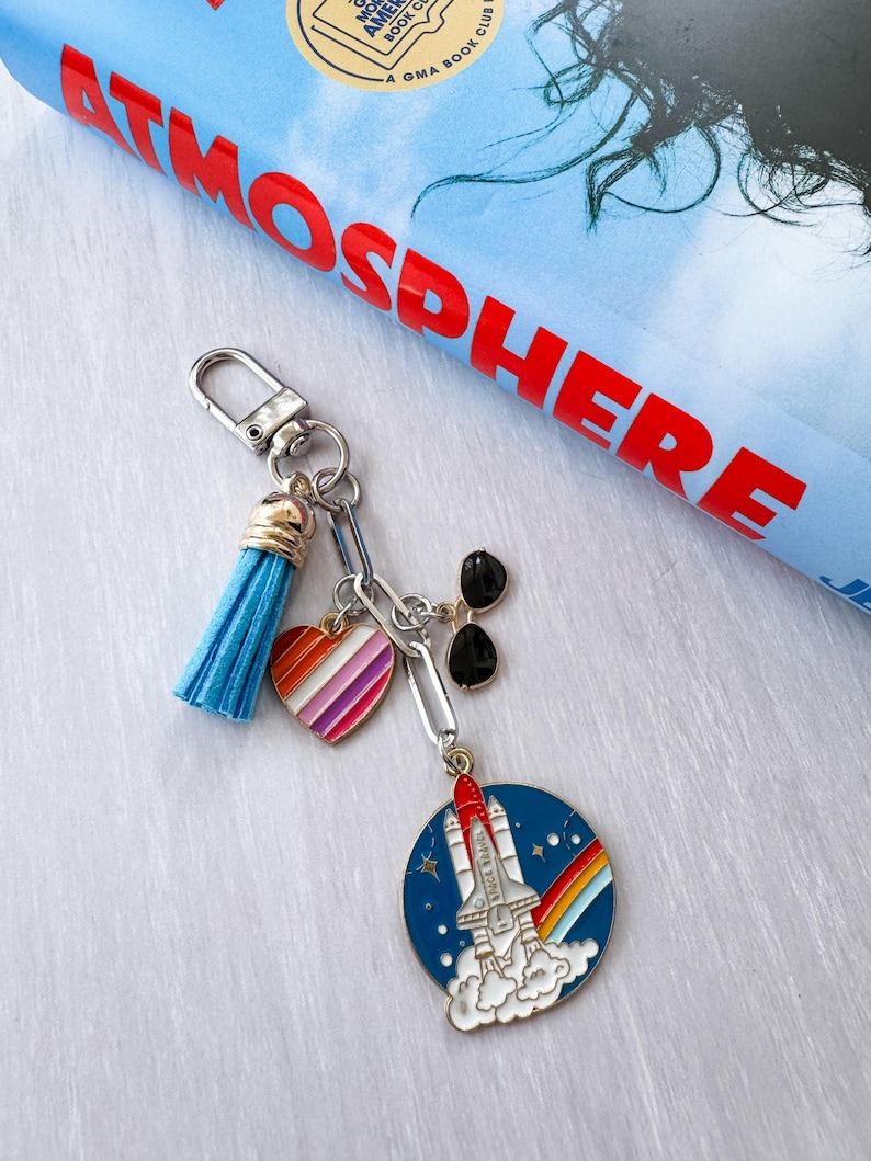 Op de afbeelding: Een zilveren sleutelhanger met een blauwe kwast, een hartvormige bedel met regenboogstrepen en een bedel met een zwarte zonnebril. De hoofdbedel is een blauwe cirkel met een raket, regenboog en sterren.