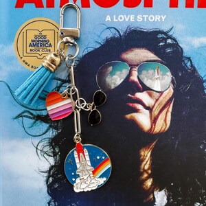 Op de afbeelding: Een zilveren sleutelhanger met een blauwe kwast, een hartbedel met regenboogstrepen en een ronde bedel met een raket en een regenboog. De achtergrond is een boekomslag met de titel "ATMOSPHERE" en de woorden "A LOVE STORY".