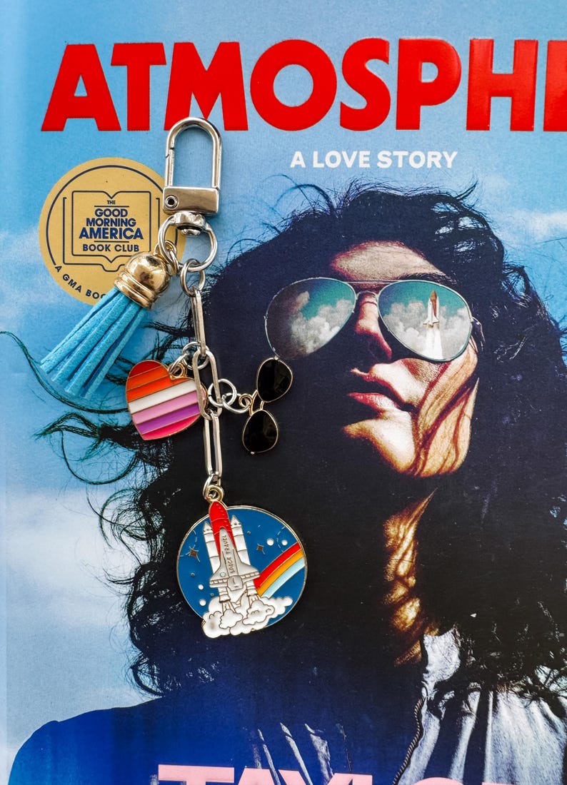 Op de afbeelding: Een zilveren sleutelhanger met een blauwe kwast, een hartbedel met regenboogstrepen en een ronde bedel met een raket en een regenboog. De achtergrond is een boekomslag met de titel "ATMOSPHERE" en de woorden "A LOVE STORY".