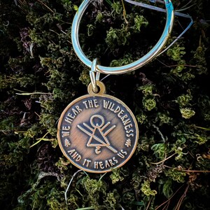 May include: A bronze pendant with the inscription "WE HEAR THE WILDERNESS AND IT HEARS US" and a geometric design, attached to a silver keyring. The background is a close-up of green moss.