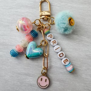 May include: A gold-toned keychain with various charms. Charms include a pink smiley face, a blue heart, a fluffy blue pom-pom, and beads spelling "PRIDE". The keychain also features a gold clasp and a chain.
