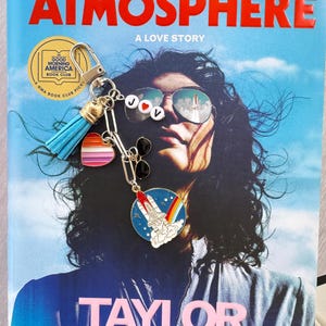 Op de afbeelding: Een boek getiteld "ATMOSPHERE: A LOVE STORY" van Taylor Jenkins Reid, met een Good Morning America Book Club-sticker en een sleutelhanger. De sleutelhanger heeft een blauwe kwast, een hart en een raketcharm.