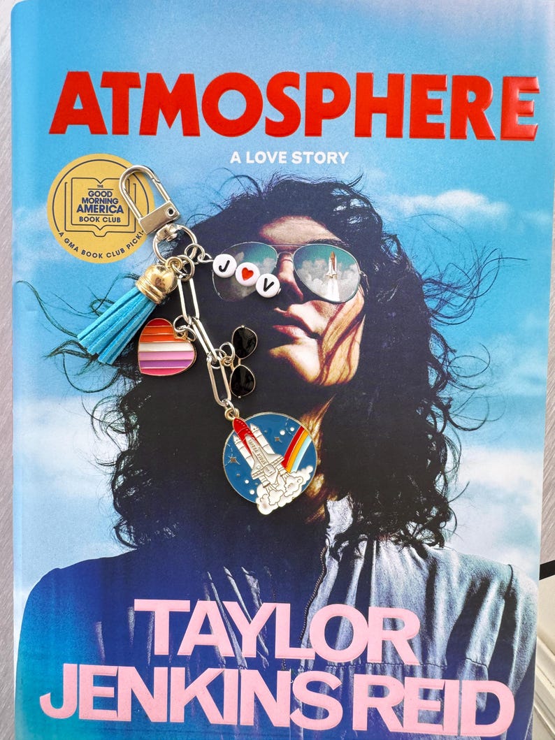 Op de afbeelding: Een boek getiteld "ATMOSPHERE: A LOVE STORY" van Taylor Jenkins Reid, met een Good Morning America Book Club-sticker en een sleutelhanger. De sleutelhanger heeft een blauwe kwast, een hart en een raketcharm.