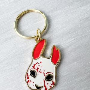 May include: A white and red enamel keychain featuring a rabbit design with black eyes and a gold-colored outline. The rabbit's ears are red, and the face has red splatter details. The keychain has a gold-colored ring.