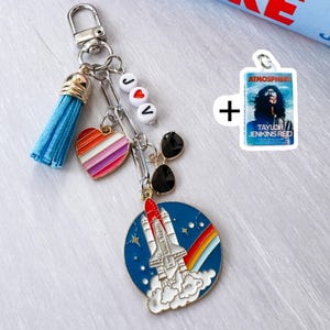 Op de afbeelding: Een zilveren sleutelhanger met een blauwe kwast, een hart met regenboogstrepen en een bedel met een raketontwerp. De sleutelhanger bevat ook een bedel met de tekst "Atmosphere" en "Taylor Jenkins Red".