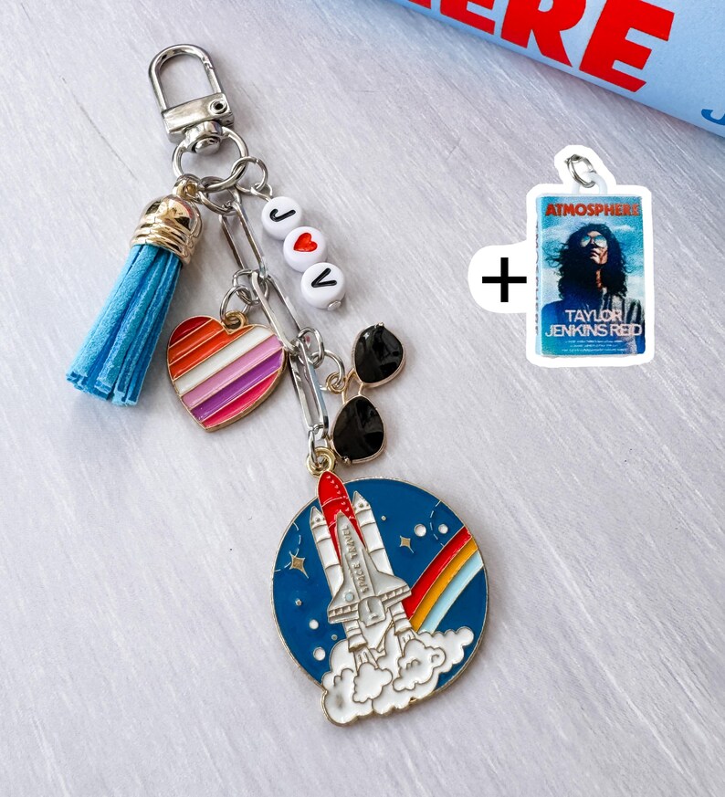 Op de afbeelding: Een zilveren sleutelhanger met een blauwe kwast, een hart met regenboogstrepen en een bedel met een raketontwerp. De sleutelhanger bevat ook een bedel met de tekst "Atmosphere" en "Taylor Jenkins Red".