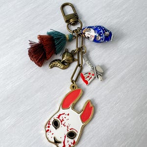 Può includere: Un portachiavi unico con un ciondolo a forma di coniglio bianco e rosso con schizzi di sangue, una bambola russa, una piccola ascia, un ciondolo a forma di volpe e un nappa. Il portachiavi è attaccato a un moschettone e una catena color bronzo.