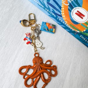 May include: An orange octopus keyring with various charms, including a cat, a horse, and a miniature book. The book reads "Remarkably Bright Creatures". A gold ring and clasp complete the keyring.