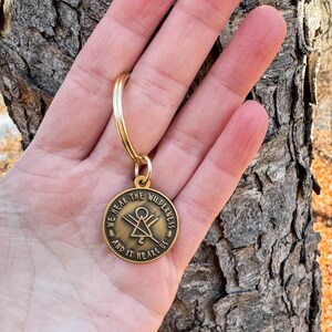 May include: A gold-coloured keyring with a circular pendant. The pendant has the text "WE HEAR THE WILDERNESS AND IT HEARS US" around a central geometric design. The keyring is held against a textured tree bark background.