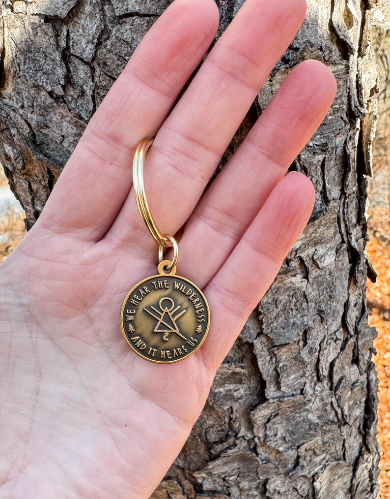 May include: A gold-coloured keyring with a circular pendant. The pendant has the text "WE HEAR THE WILDERNESS AND IT HEARS US" around a central geometric design. The keyring is held against a textured tree bark background.