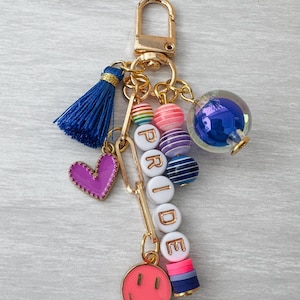 Puede incluir: Un llavero dorado con una borla azul, un corazón morado y un dije de carita sonriente rosa. El llavero presenta cuentas con la palabra "PRIDE", cuentas a rayas arcoíris, rosas y azules, y un globo de cristal azul.
