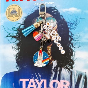 Op de afbeelding: Een boekomslag met de titel "ATMOSPHERE" en auteur "TAYLOR JENKINS REID" wordt getoond. Een sleutelhanger met een blauwe kwast, een hart met regenboogstrepen en een ruimteveer-bedel hangt voor het boek. De sleutelhanger heeft ook de woorden "WE ARE THE STARS".