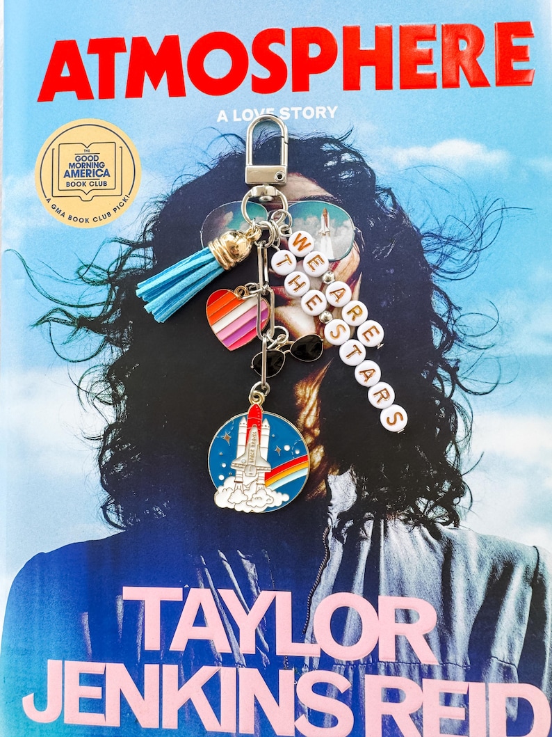 Op de afbeelding: Een boekomslag met de titel "ATMOSPHERE" en auteur "TAYLOR JENKINS REID" wordt getoond. Een sleutelhanger met een blauwe kwast, een hart met regenboogstrepen en een ruimteveer-bedel hangt voor het boek. De sleutelhanger heeft ook de woorden "WE ARE THE STARS".