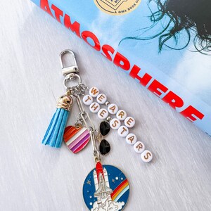 Op de afbeelding: Een zilveren sleutelhanger met meerdere bedels, waaronder een blauwe kwast, een hart met regenboogstrepen en een ronde bedel met een raketontwerp. De sleutelhanger heeft ook de tekst "WE ARE THE STARS" uitgespeld in witte letterkralen.
