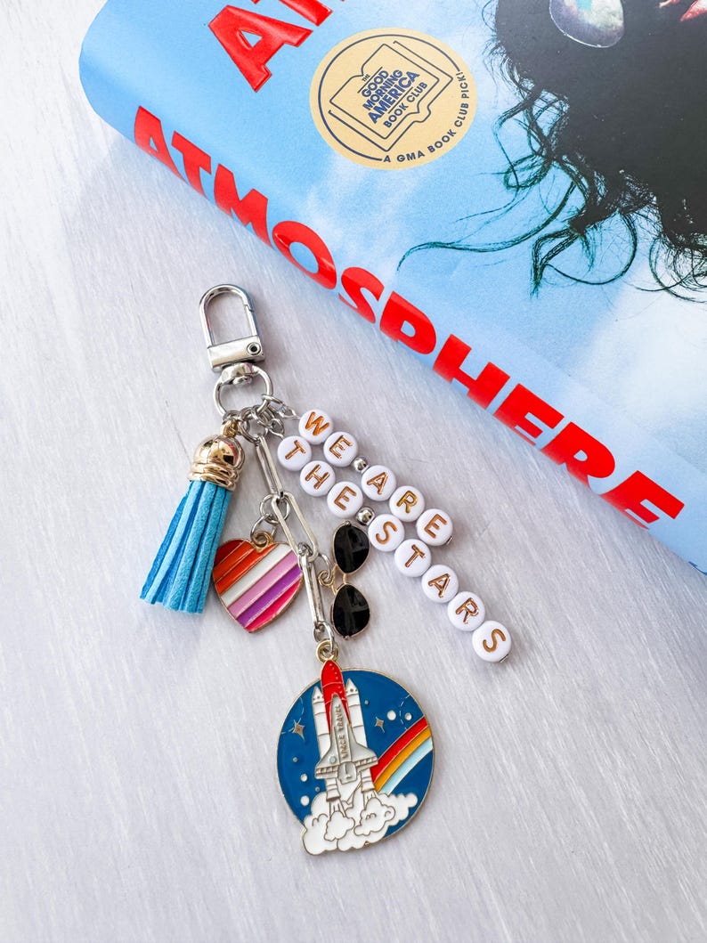 Op de afbeelding: Een zilveren sleutelhanger met meerdere bedels, waaronder een blauwe kwast, een hart met regenboogstrepen en een ronde bedel met een raketontwerp. De sleutelhanger heeft ook de tekst "WE ARE THE STARS" uitgespeld in witte letterkralen.