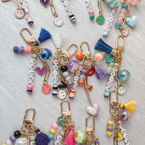 May include: A collection of colorful keychains with gold-tone hardware. Each keychain features beaded accents, tassels, and charms, including smiley faces, hearts, and the word "PRIDE" spelled out with letter beads. The keychains are designed with a variety of colors.