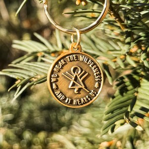 May include: A gold-coloured keyring with a circular pendant. The pendant features the text "WE HEAR THE WILDERNESS AND IT HEARS US". A symbol is in the centre. The keyring hangs against a blurred green backdrop.