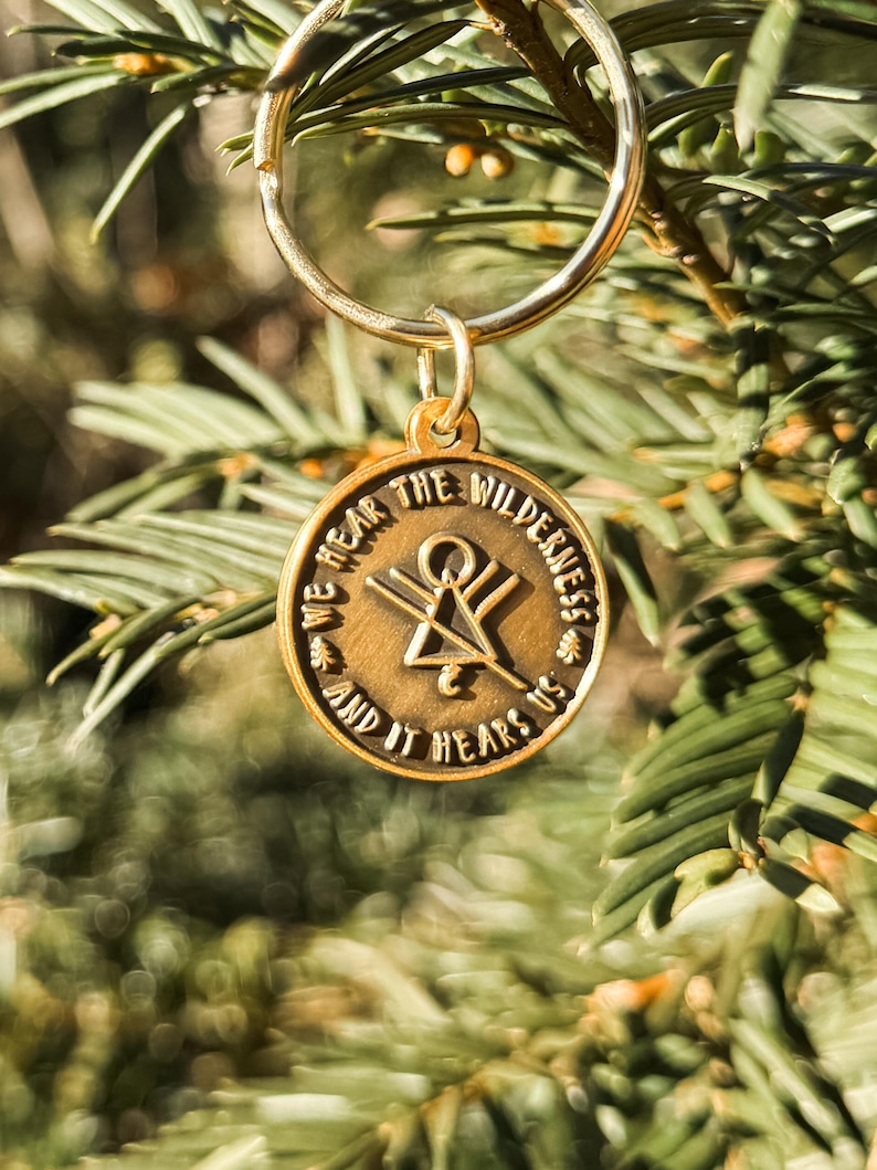 May include: A gold-coloured keyring with a circular pendant. The pendant features the text "WE HEAR THE WILDERNESS AND IT HEARS US". A symbol is in the centre. The keyring hangs against a blurred green backdrop.