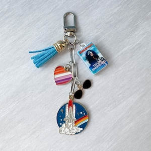 Op de afbeelding: Een zilveren sleutelhanger met een blauwe kwast, een kleine boekenbedel, een hartbedel met regenboogstrepen, een zwarte zonnebril en een ronde bedel met een spaceshuttle-ontwerp en een regenboog. De boekenbedel leest "Atmosphere" van Taylor Jenkins Reid.