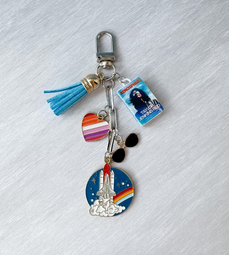 Op de afbeelding: Een zilveren sleutelhanger met een blauwe kwast, een kleine boekenbedel, een hartbedel met regenboogstrepen, een zwarte zonnebril en een ronde bedel met een spaceshuttle-ontwerp en een regenboog. De boekenbedel leest "Atmosphere" van Taylor Jenkins Reid.