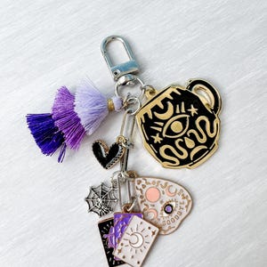 May include: A keychain with a silver clasp and multiple charms. Charms include a black and gold cup with an eye design, a black heart, a spiderweb, and tarot card charms. A purple and gold tassel hangs from the clasp.