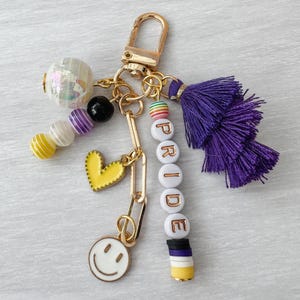 May include: A gold-tone keychain with a variety of charms. It features a purple tassel, beads spelling "PRIDE", a yellow heart, a smiley face, and other colorful beads. The keychain is designed to be attached to keys or a bag.