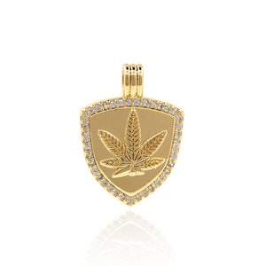 Op de afbeelding: Goudkleurige hanger met een schildvorm, met een cannabisbladontwerp. De hanger is omrand met kleine, heldere edelstenen. De beugel is een eenvoudig lusontwerp. De hanger is waarschijnlijk gemaakt van verguld metaal.