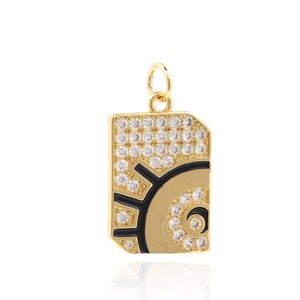 Puede incluir: Colgante dorado con forma rectangular, con un diseño de sol u ojo con detalles en negro y beige. La mitad superior está incrustada con pequeños cristales transparentes. Un pequeño lazo en la parte superior para una cadena.