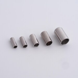 May include: Five cylindrical metal tubes of varying sizes are arranged on a white surface. The tubes are silver in color and have a smooth, polished finish. The open ends of the tubes reveal a hollow interior.