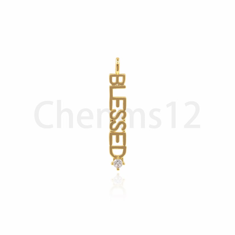 Pode incluir: Encanto de pingente de ouro com a palavra "BLESSED" escrita verticalmente e um pequeno strass transparente na parte inferior.